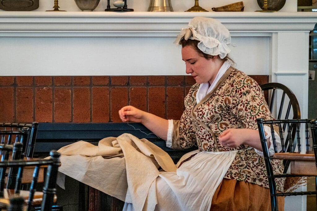 Boston, MA - US - Oct 2, 2025 A historical reenactor sewing a piece of fabric in a room with a fireplace and colonial-style furniture at the Boston Tea Party Ships and Museum in Boston